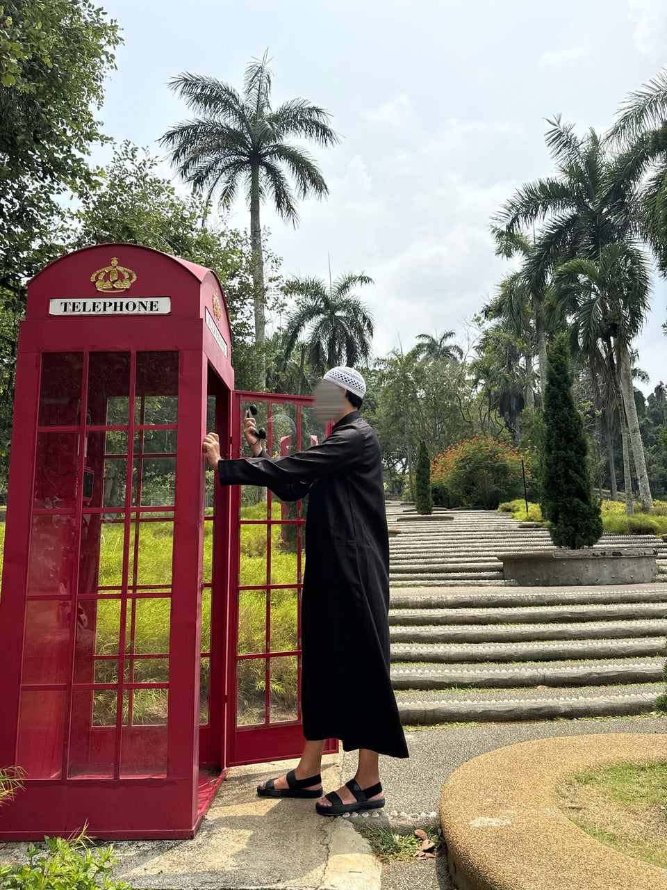 Une silhouette en qamis noir près d'une cabine téléphonique rouge et de palmiers.