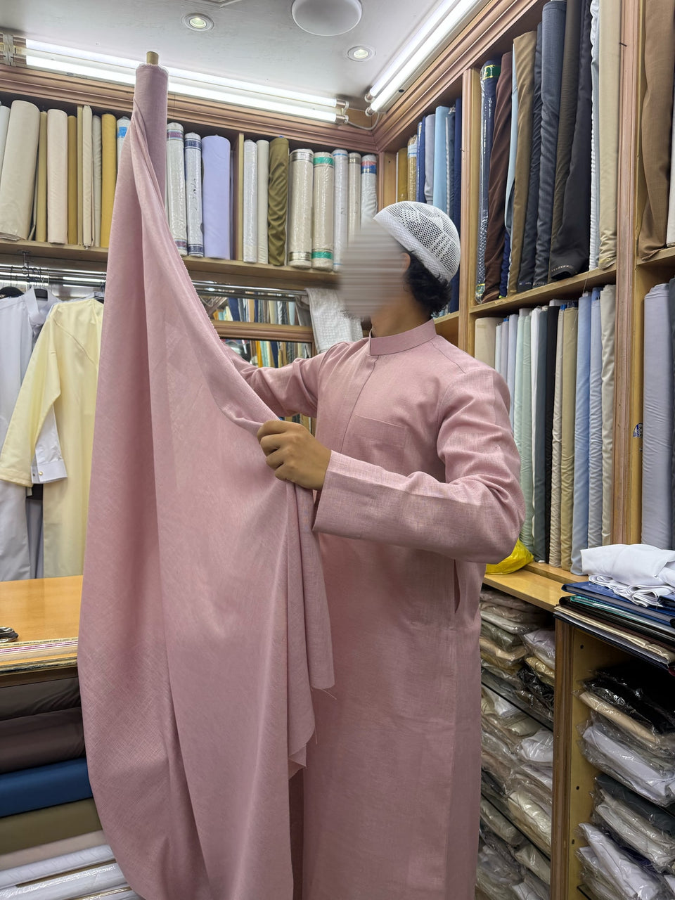 Une silhouette en qamis rose tenant un tissu rose parmi des étoffes colorées.