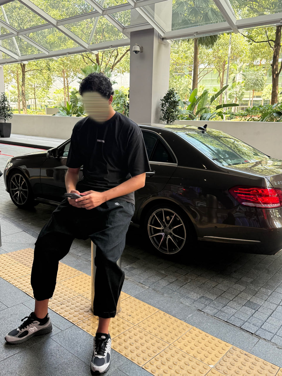Une tenue noire décontractée, assis devant une voiture, téléphone à la main.